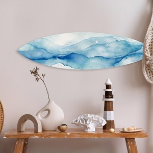 May include: A surfboard-shaped wall art piece with a watercolor design in shades of blue and white hangs on a light-colored wall. A wooden table displays decorative items, including a lighthouse and a vase.