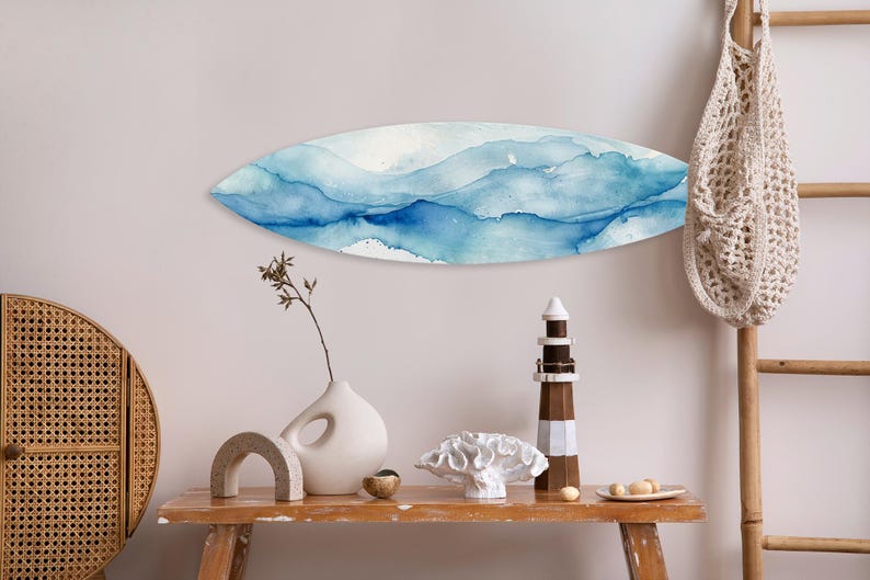 May include: A surfboard-shaped wall art piece with a watercolor design in shades of blue and white hangs on a light-colored wall. A wooden table displays decorative items, including a lighthouse and a vase.