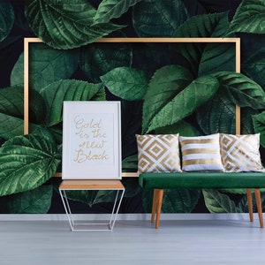 Op de afbeelding: Een groene fluwelen bank met gouden en witte gepatroneerde kussens staat voor een donkergroene en zwarte behang met grote bladeren. Een wit frame met de tekst "Gold is the new Black" staat op een witte tafel naast de bank.