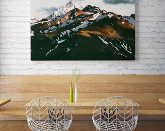 May include: A framed photograph of a mountain range with snow-capped peaks and green slopes. The photograph is hanging on a white brick wall above a wooden table with two white metal chairs.