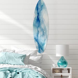 May include: A surfboard with a blue watercolor design hangs on a white shiplap wall. The surfboard has a teardrop shape with shades of blue and white. The room has a coastal theme, with a bed, pillows, and a turquoise lamp.