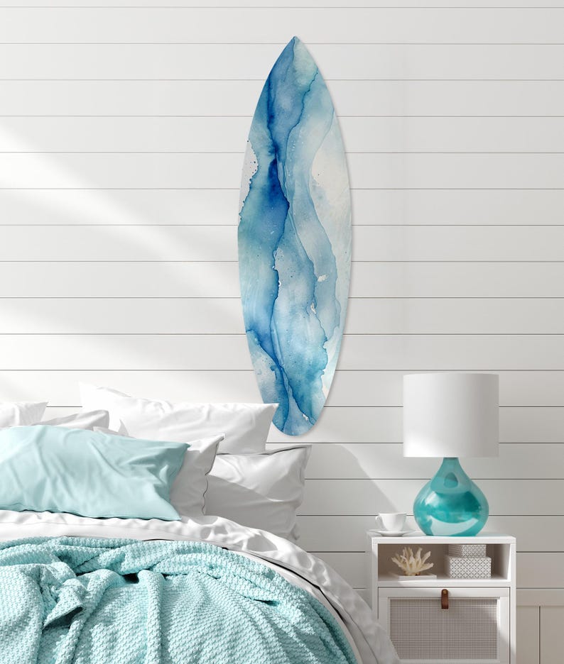 May include: A surfboard with a blue watercolor design hangs on a white shiplap wall. The surfboard has a teardrop shape with shades of blue and white. The room has a coastal theme, with a bed, pillows, and a turquoise lamp.