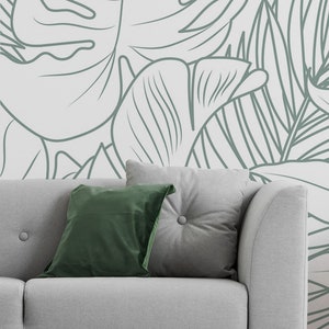 Puede incluir: Un sof&aacute; gris con dos almohadas, una verde y una gris, se encuentra frente a una pared con un patr&oacute;n de hojas tropicales blancas y verdes.