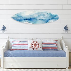 May include: A coastal-themed bedroom with a white daybed, blue bedding, and striped pillows. A surfboard-shaped wall art with blue watercolor design hangs above the bed. Nautical decor includes a pillow with a ship's wheel design.