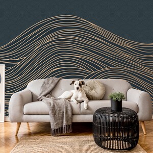 May include: A white dog sits on a light gray couch in front of a dark blue wall with a gold abstract wave pattern. The couch has a gray throw blanket and a beige pillow. A black side table with a potted plant sits in front of the couch.