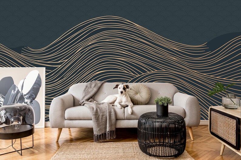 May include: A white dog sits on a light gray couch in front of a dark blue wall with a gold abstract wave pattern. The couch has a gray throw blanket and a beige pillow. A black side table with a potted plant sits in front of the couch.