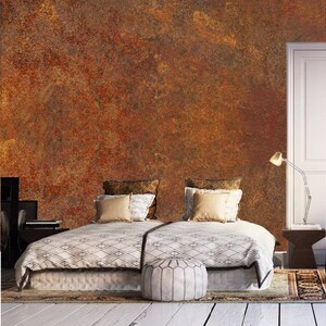 May include: A bedroom with a bed with a white and black patterned duvet, two pillows, and a white ottoman. The wall behind the bed is painted with a rusty orange and brown textured paint. There is a lamp on a small table next to the bed.
