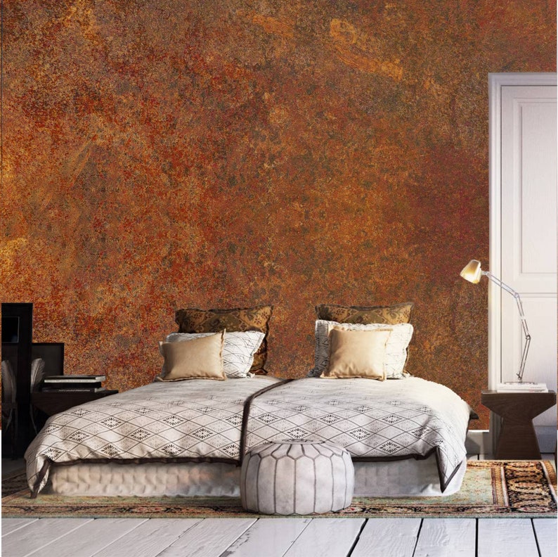 May include: A bedroom with a bed with a white and black patterned duvet, two pillows, and a white ottoman. The wall behind the bed is painted with a rusty orange and brown textured paint. There is a lamp on a small table next to the bed.