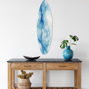 May include: A surfboard-shaped wall art piece with a watercolor design in shades of blue and white. The art is mounted on a white wall above a wooden console table. A blue vase with a plant and a decorative bowl are also on the table.