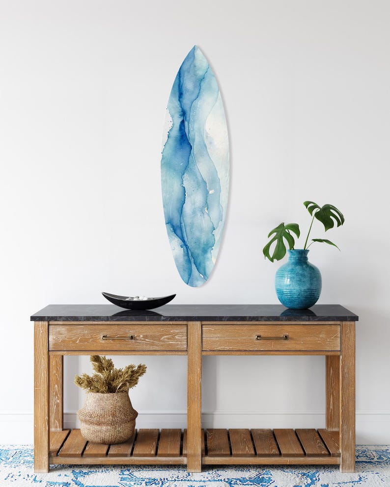 May include: A surfboard-shaped wall art piece with a watercolor design in shades of blue and white. The art is mounted on a white wall above a wooden console table. A blue vase with a plant and a decorative bowl are also on the table.