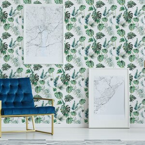 Puede incluir: Un sillón de terciopelo azul teal con un marco de metal dorado se encuentra frente a una pared blanca con un papel tapiz de estampado de hojas tropicales. Dos mapas enmarcados blancos cuelgan de la pared.