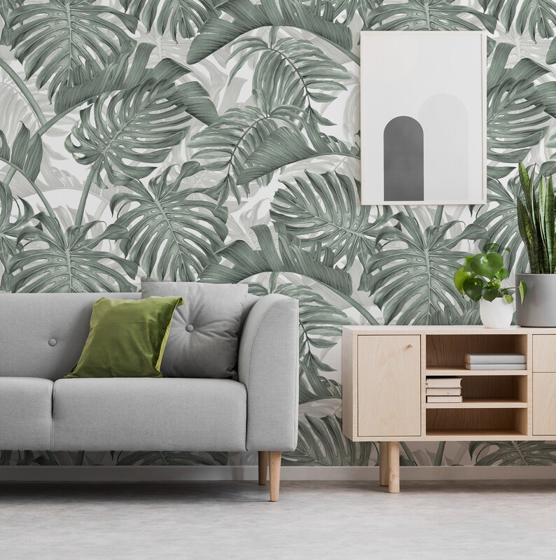 May include: A gray sofa with two green throw pillows sits in front of a wall with a tropical leaf wallpaper pattern. A wooden cabinet with shelves is to the right of the sofa. A potted plant is on the cabinet.