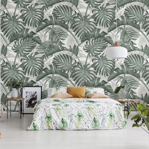 May include: A bedroom with a white wall featuring a green tropical leaf wallpaper. A bed with a white and green floral duvet cover is in the center of the room. A wicker chair with a brown throw blanket is in the left corner of the room. A small table with a potted plant is in the right corner of the room.
