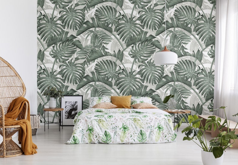 May include: A bedroom with a white wall featuring a green tropical leaf wallpaper. A bed with a white and green floral duvet cover is in the center of the room. A wicker chair with a brown throw blanket is in the left corner of the room. A small table with a potted plant is in the right corner of the room.