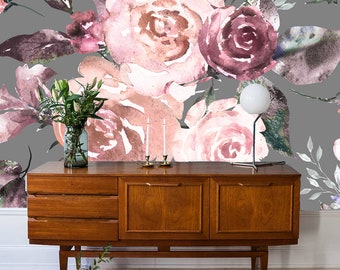 May include: A gray wall with a floral wallpaper featuring pink and burgundy watercolor roses and leaves. A wooden dresser with three drawers and two doors sits in front of the wall. A small table with a vase of greenery and candles sits in front of the dresser.