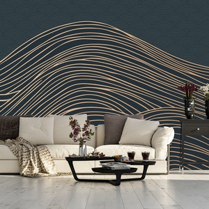 May include: A living room interior design with a dark blue wall featuring a gold and white abstract wave pattern wallpaper. A white sofa with a throw blanket sits in front of the wall. A black coffee table with a glass top sits in front of the sofa. A floor lamp with a blue lampshade stands to the left of the sofa. A black console table with three drawers sits to the right of the sofa. The console table has a vase with white flowers and a vase with purple flowers on top.