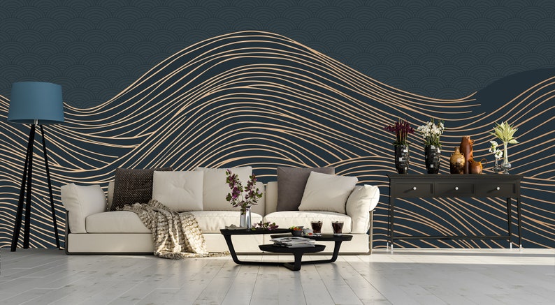 May include: A living room interior design with a dark blue wall featuring a gold and white abstract wave pattern wallpaper. A white sofa with a throw blanket sits in front of the wall. A black coffee table with a glass top sits in front of the sofa. A floor lamp with a blue lampshade stands to the left of the sofa. A black console table with three drawers sits to the right of the sofa. The console table has a vase with white flowers and a vase with purple flowers on top.