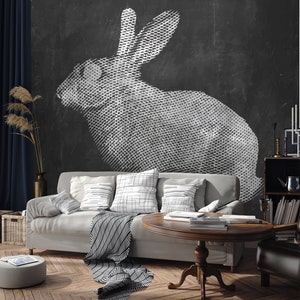 May include: A white rabbit silhouette drawn in a stippled style on a black chalkboard wall. The rabbit is facing left and is in a sitting position. The wall is behind a light gray sofa with black and white patterned throw pillows and a black and white patterned throw blanket.