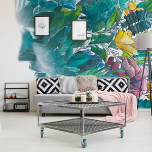 May include: A modern living room with a gray sofa, decorative pillows, and a pink throw blanket. A metal coffee table with wheels sits in front of the sofa. The wall features a large mural with a face and floral design.