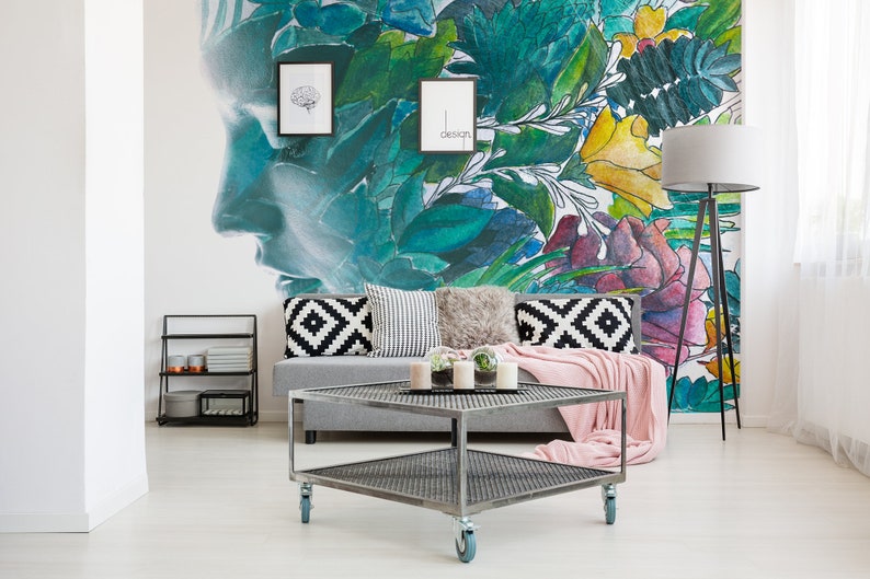 May include: A modern living room with a gray sofa, decorative pillows, and a pink throw blanket. A metal coffee table with wheels sits in front of the sofa. The wall features a large mural with a face and floral design.