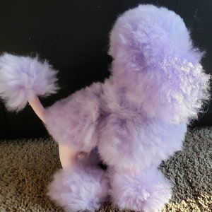 K&ouml;nnte beinhalten: Ein Pl&uuml;sch-Pudel-Spielzeug in Lavendel mit einem flauschigen Schwanz und einer rosa Nase.
