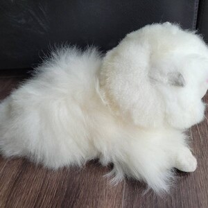 May include: A white fluffy plush toy resembling a kitten or a small cat. The toy has a soft, furry texture and is lying on a wooden surface.