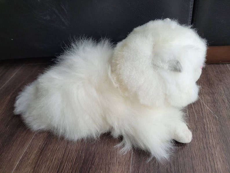 May include: A white fluffy plush toy resembling a kitten or a small cat. The toy has a soft, furry texture and is lying on a wooden surface.