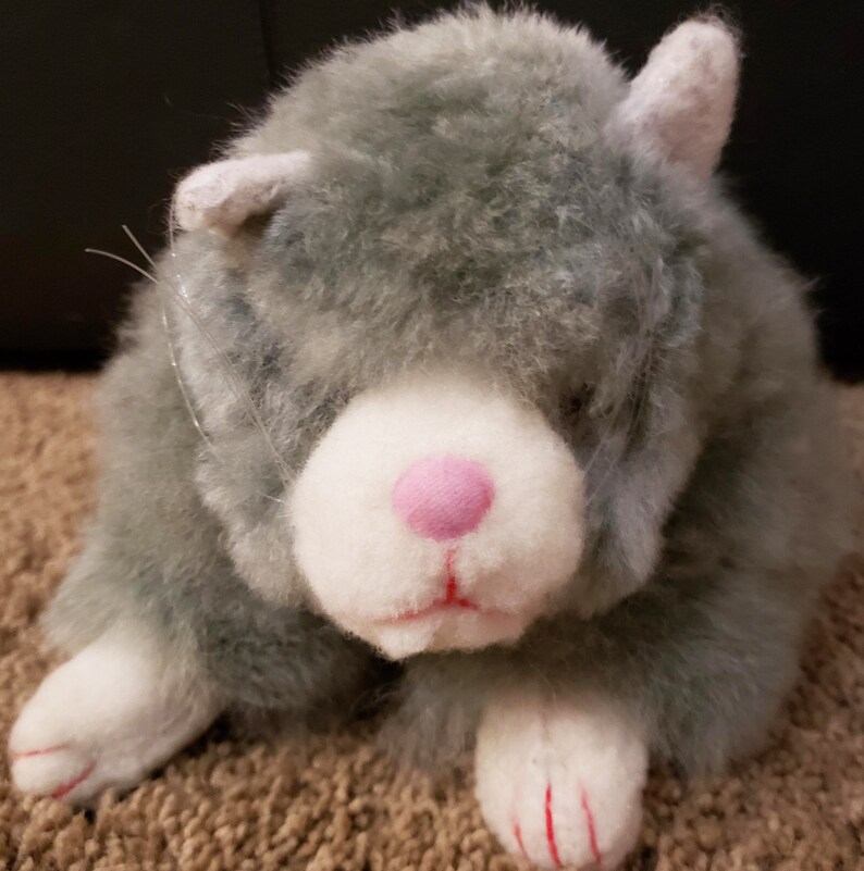 May include: A gray and white plush cat toy with pink nose and red claws. The cat is sitting on a brown carpet.
