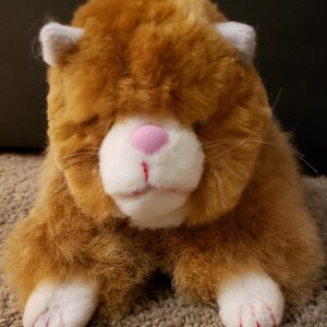 May include: A plush orange and white cat toy with pink nose and red claws. The cat is lying down with its head resting on its paws.