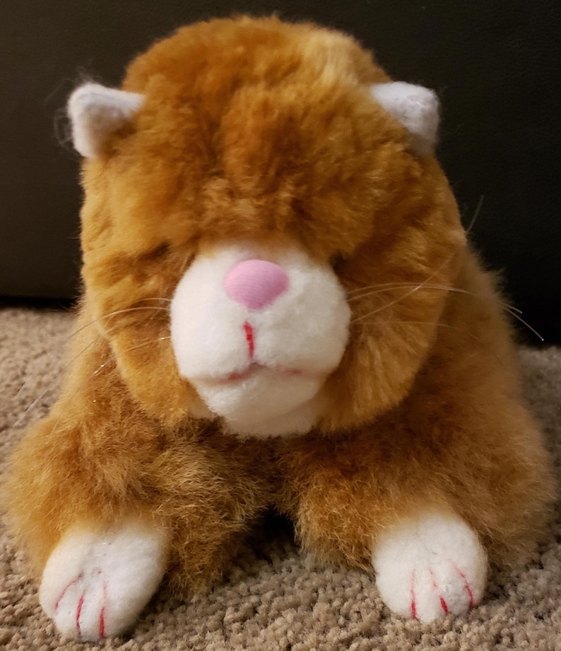 May include: A plush orange and white cat toy with pink nose and red claws. The cat is lying down with its head resting on its paws.