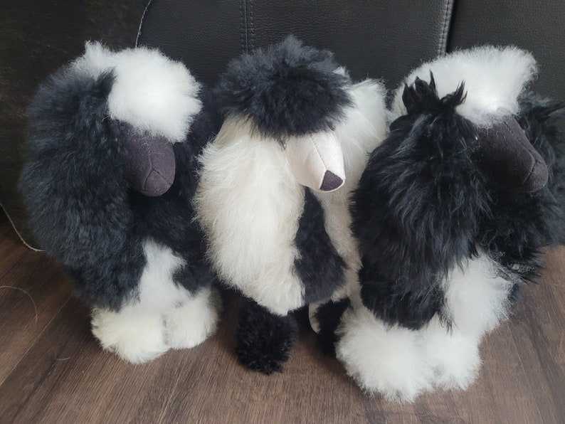 K&ouml;nnte beinhalten: Drei schwarze und wei&szlig;e flauschige Alpaka-Pl&uuml;schtiere. Die Pl&uuml;schtiere stehen auf einem Holzboden.