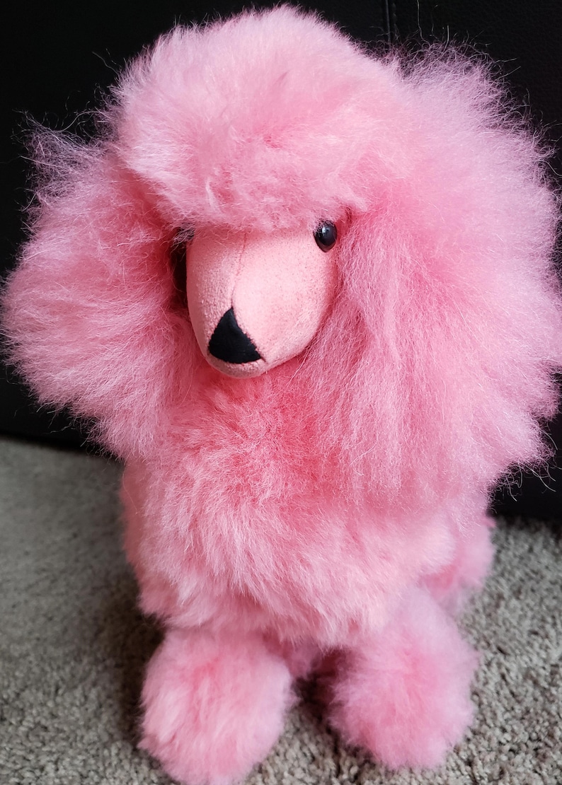 K&ouml;nnte beinhalten: Ein flauschiges rosa Pl&uuml;schtier in Form eines Pudels. Das Spielzeug hat eine schwarze Nase und rosa Augen.