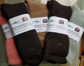 May include: Four pairs of alpaca socks in different colors, including pink, brown, black, and gray. The socks are packaged with labels that read "Homegrown Alpaca Socks" and include the website address "AuroraAlpacasLlamas.com".