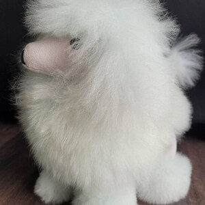 K&ouml;nnte beinhalten: Ein wei&szlig;er, flauschiger Spielzeugpudel mit rosa Nase und schwarzen Augen. Das Spielzeug steht auf einer Holzoberfl&auml;che.