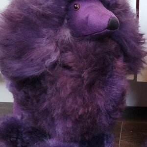 K&ouml;nnte beinhalten: Ein Pl&uuml;schtier in Form eines lila Pudels mit flauschigem Fell. Das Spielzeug hat ein braunes Auge und eine schwarze Nase.