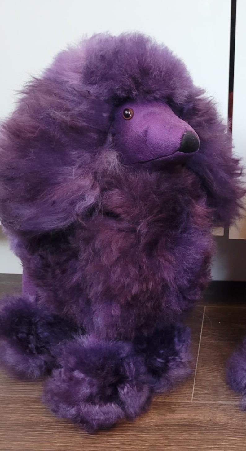 K&ouml;nnte beinhalten: Ein Pl&uuml;schtier in Form eines lila Pudels mit flauschigem Fell. Das Spielzeug hat ein braunes Auge und eine schwarze Nase.