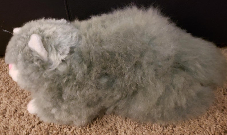 May include: A plush gray cat toy with white fur accents around the face and paws. The toy is made of soft, fluffy material and has a realistic cat appearance.