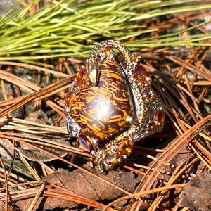May include: A detailed close-up of a statement ring featuring a large, oval-shaped stone with a unique brown and orange pattern. The ring is adorned with smaller orange gemstones and intricate metalwork, creating a luxurious and eye-catching design.