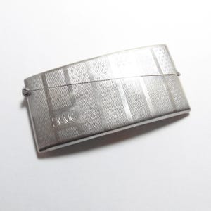 May include: A rectangular, silver-toned metal case with a hinged lid. The case features an engraved pattern of vertical stripes and floral designs. The initials "J.R.C." are engraved on the front. The case is slightly curved and has a clasp closure.