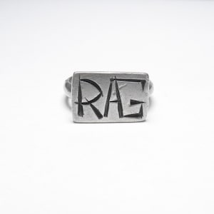 May include: A silver ring with a rectangular face. The face of the ring has the letters "RAG" etched into it. The ring is set against a white background.