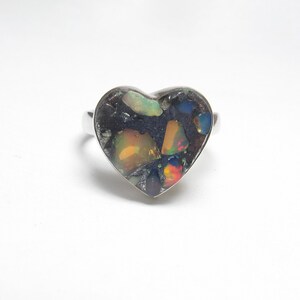 May include: A heart-shaped ring featuring a dark, textured inlay embedded with multiple iridescent opal fragments. The colorful opal pieces display flashes of orange, green, and blue against a dark background, all set within a polished silver-toned metal band.