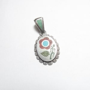 May include: A silver pendant featuring a turquoise and red flower design with a stylised human figure. The pendant is surrounded by a silver border with a scalloped edge.