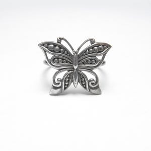 May include: A silver-toned butterfly ring. The ring features a detailed butterfly design with openwork wings, dotted accents, and a curved band. The butterfly's body is a solid piece, and the wings have a delicate, airy appearance.