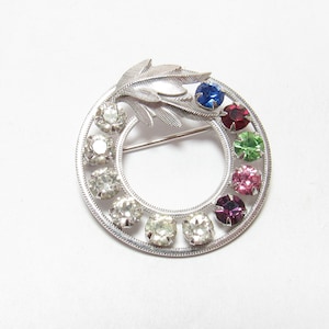 May include: A silver brooch with a leaf design and a circle of clear and colored rhinestones. The rhinestones are arranged in a rainbow pattern.