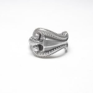 May include: A silver spoon ring with an ornate design. The ring is made from a bent spoon and has a decorative pattern.