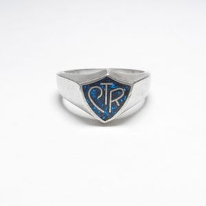 May include: A silver-toned ring featuring a shield-shaped face with a dark blue, glittery inlay. The letters CTR are prominently displayed in silver on the blue background. The band is smooth and tapers toward the back of the ring.