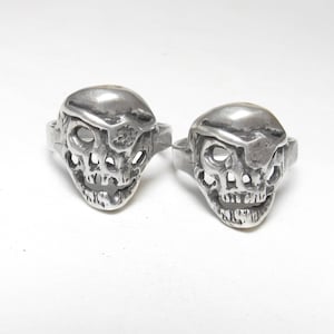 Może przedstawiać: Dwa srebrne pierścienie w kształcie czaszki z wzorem kapelusza pirata. Pierścienie są ozdobione oczodołami, nosem i ustami.