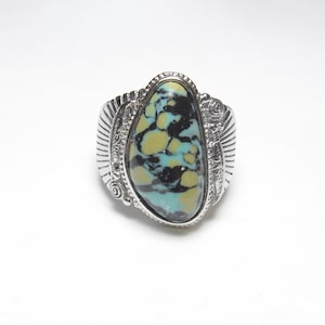 May include: A silver ring with an oval turquoise stone, displaying a pattern of black, yellow, and blue-green. The ring has a detailed, textured band with decorative elements.
