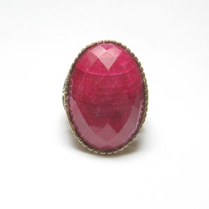 May include: A ring featuring a large, oval-shaped, faceted, pink agate stone set in a gold-toned metal bezel.