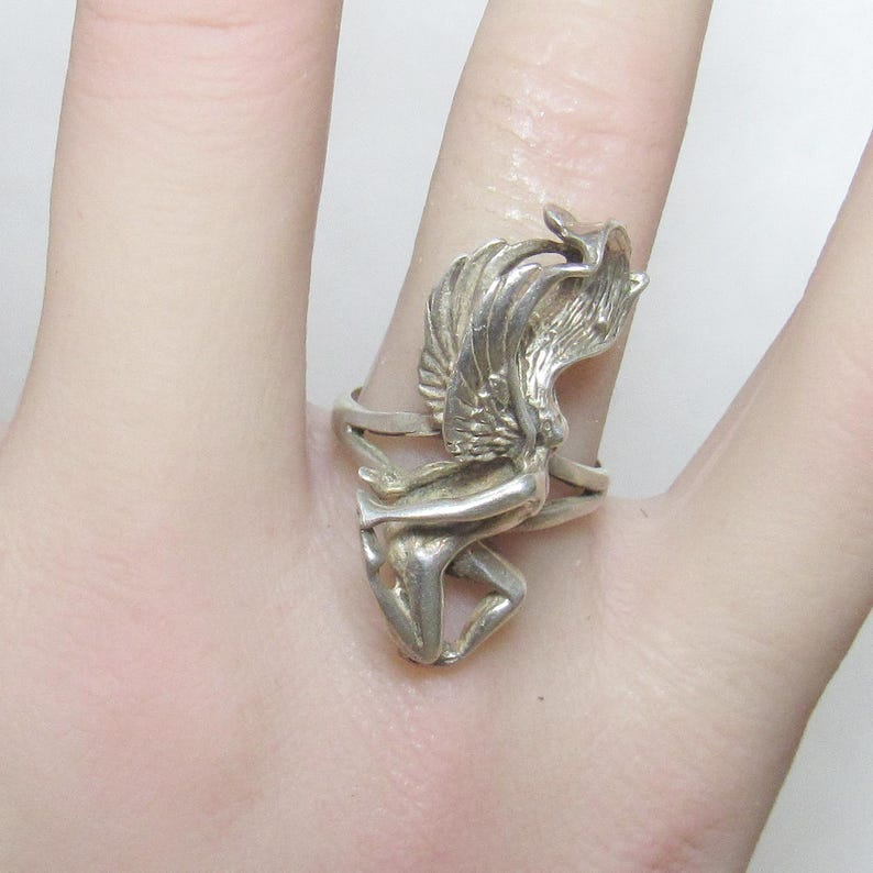 Puede incluir: Un anillo de plata con una figura alada esculpida. El dise&ntilde;o del anillo incluye una representaci&oacute;n detallada de una persona con alas extendidas, creando una joya &uacute;nica y art&iacute;stica. El anillo se lleva en un dedo.